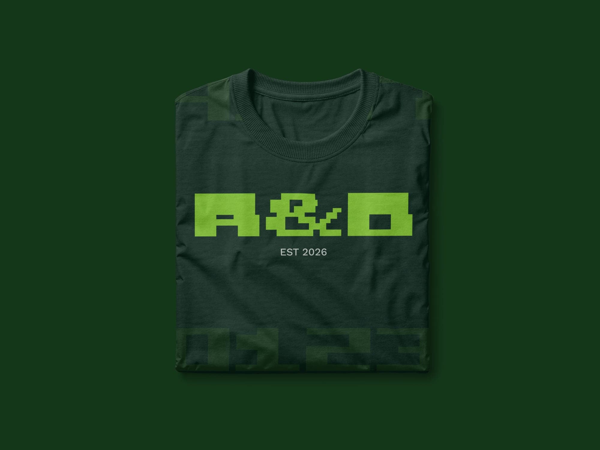 Folded dark green T-shirt on green, printed with A&D in lime Riteblock above EST 2026, with large faded letters cropped at its edges.